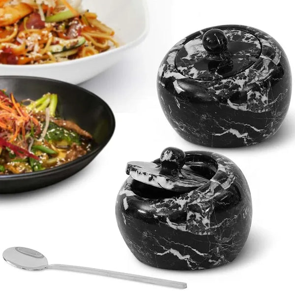 Two 4 inch black marble salt cellars with lids and a stainless steel spoon beside plated noodle dishes