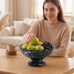 Woman placing a green apple into a black marble pedestal fruit bowl full of fruit on a coffee table