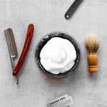 Black marble shaving bowl with lather flanked by razors, brush and towel on white floor