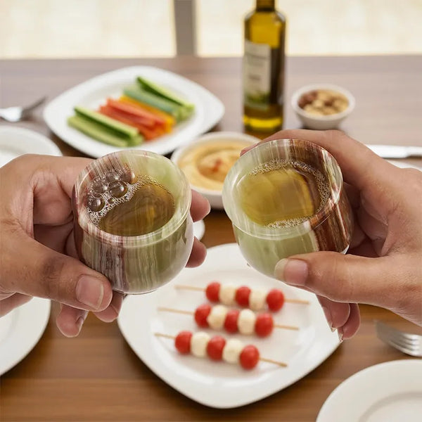 Two hands toasting with green onyx marble shot glasses over a table of appetizers