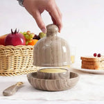 Hand lifting the dome lid off a natural brown marble butter dish set on a breakfast table with fruit and toast