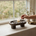 Hand lighting a candle on an oceanic footed marble tray with pebbles by a window