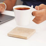 Square travertine coaster on a desk as a hand lifts a white coffee mug