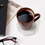 White octagon marble coaster under a white coffee cup beside a newspaper and glasses