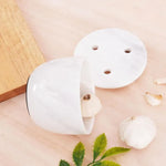 White marble garlic keeper tipped on its side with the vented lid off, showing the felt base ring, beside fresh garlic