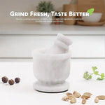 White marble mortar and pestle on a kitchen counter with whole peppercorns, cardamom pods and fresh coriander