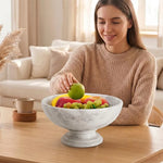 Woman placing a green apple into a white marble pedestal fruit bowl full of fruit on a coffee table