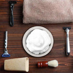 White marble shaving bowl with lather flanked by razors, brush and towel on wood