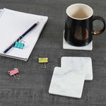 Square white marble coasters beside a black mug and notepad on a desk