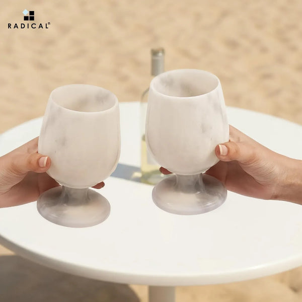 Two hands toasting white marble wine glasses at an outdoor table with white wine