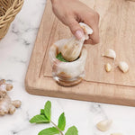 Hand grinding fresh green herbs into a paste with a white onyx marble mortar and pestle, beside garlic, ginger and mint
