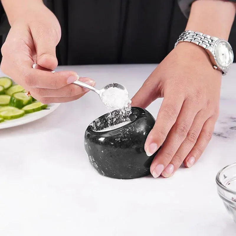 Spoon lifting pink salt from a black marble salt cellar held steady on a countertop, 3 inches wide and tall