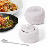 Two 4 inch white marble salt cellars with lids and a stainless steel spoon beside plated noodle dishes