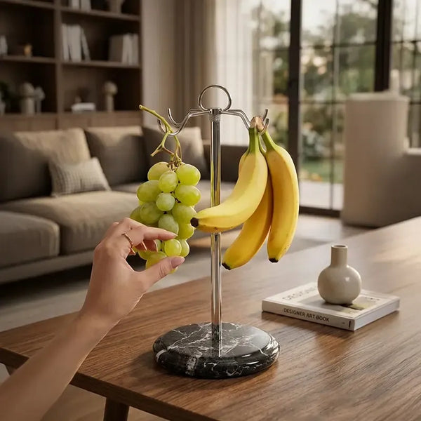 Black marble banana holder on a wooden table holding bananas and grapes, hand reaching for the grapes