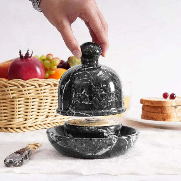 Hand lifting the dome lid off a black marble butter dish set on a breakfast table with fruit and toast