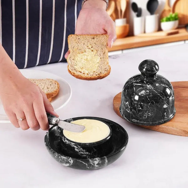 Butter being spread on toast with the marble knife from a black marble butter dish, dome lid resting on a wooden board