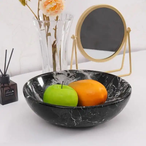 Black marble fruit bowl holding a green apple and a mango on a console table beside a vase and mirror