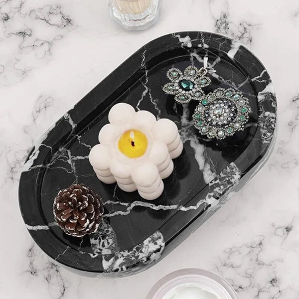 Black marble oval tray holding a candle, earrings and a pinecone on marble