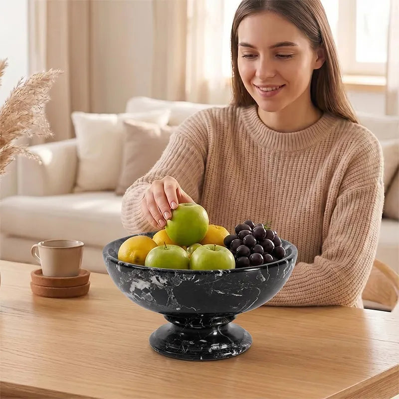 Woman placing a green apple into a black marble pedestal fruit bowl full of fruit on a coffee table