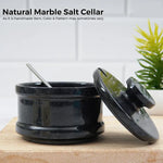 Black marble stone salt keeper with spoon inside and lid leaning against it on a wooden board