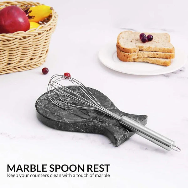 Black marble spoon rest holding a whisk on a kitchen counter with fruit and toast