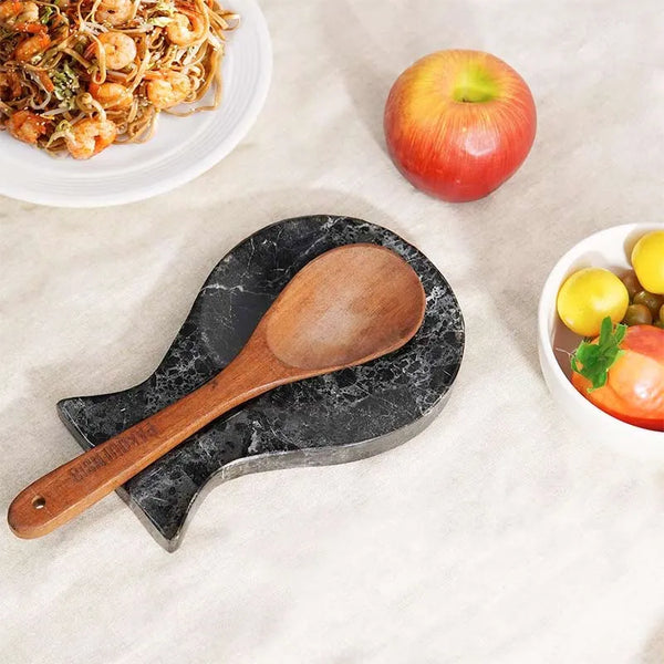 Black marble utensil rest holding a wooden spoon beside a plate of noodles