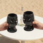 Two hands toasting black marble wine glasses at an outdoor table with white wine