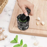 Hand grinding fresh green herbs into a paste with a black zebra marble mortar and pestle, beside garlic, ginger and mint