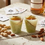 Two green onyx marble shot glasses of whiskey on a rustic table with cards and nuts