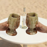 Two hands toasting green onyx marble wine glasses at an outdoor table with white wine