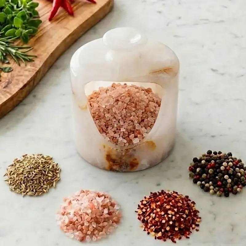 White onyx marble salt pig with an open front cut-out, surrounded by piles of pink salt, chilli flakes, cumin and peppercorns