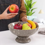 Hand placing an apple into a oceanic marble pedestal fruit bowl holding pomegranate, pear and banana