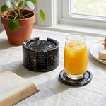 Round black marble coaster set with holder beside a glass of orange juice on a linen cloth