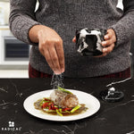 Salt being sprinkled onto a plated steak from a black marble seasoning keeper held in hand