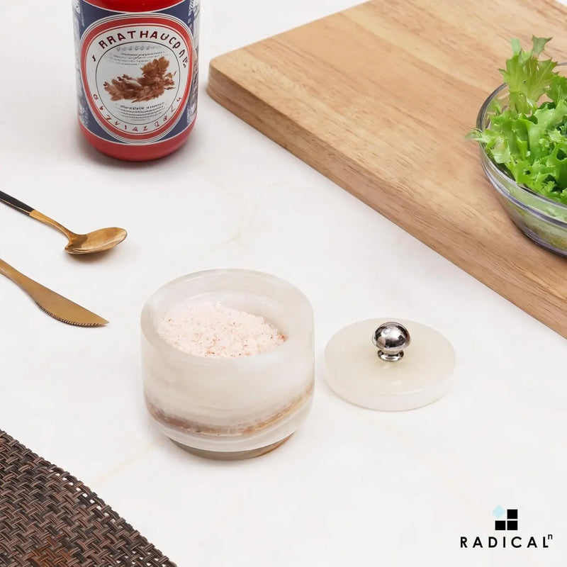 Small white onyx marble seasoning keeper filled with salt, stainless steel knob lid set to the side on a countertop