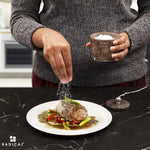 Salt being sprinkled onto a plated steak from a oceanic marble seasoning keeper held in hand