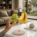 Verona marble banana holder on a white wooden table holding bananas and grapes, hand reaching for the grapes