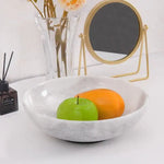 White marble fruit bowl holding a green apple and a mango on a console table beside a vase and mirror
