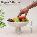 White marble pedestal fruit bowl measuring 10 inches wide and 5 inches tall, filled with fruit as a hand reaches in
