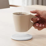 Round white marble coaster on a wooden table as a hand lifts a cup of coffee
