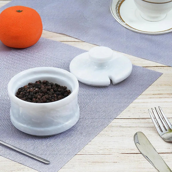 White marble salt cellar filled with peppercorns, notched lid set aside on a table with fruit and cutlery