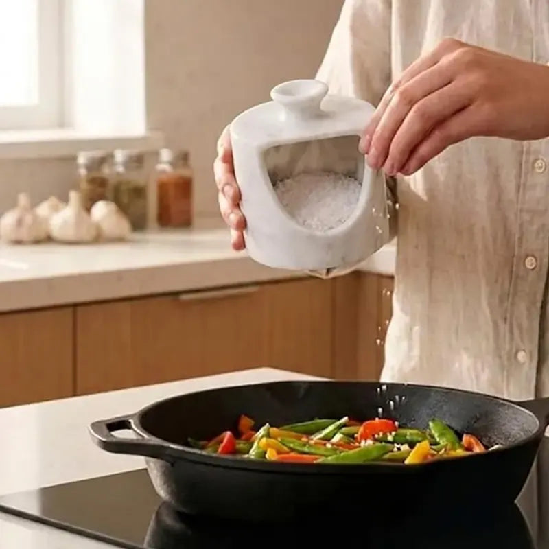 White marble salt pig held in hand pouring salt into a pan of vegetables on the stove