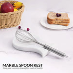 White marble spoon rest holding a whisk on a kitchen counter with fruit and toast