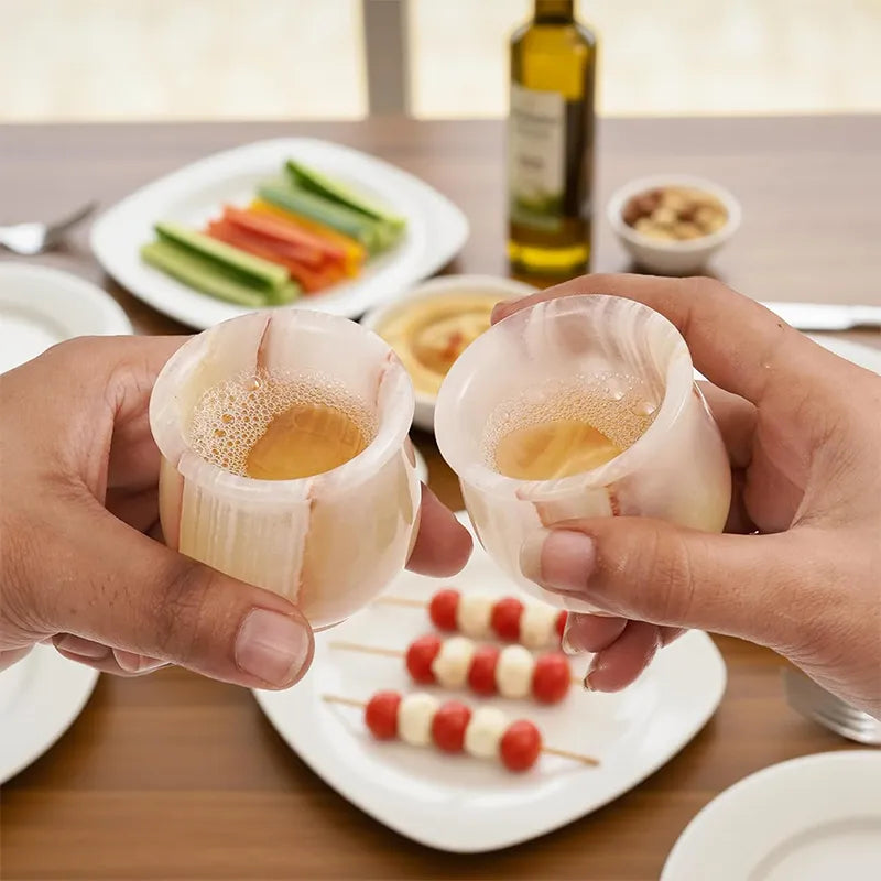 Two hands toasting with white onyx marble shot glasses over a table of appetizers