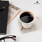 White onyx square marble coaster under a white coffee cup beside a newspaper and glasses